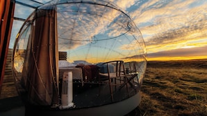 Una estructura de vidrio esférica con una cama y una mesa en su interior, sobre un cielo al atardecer.