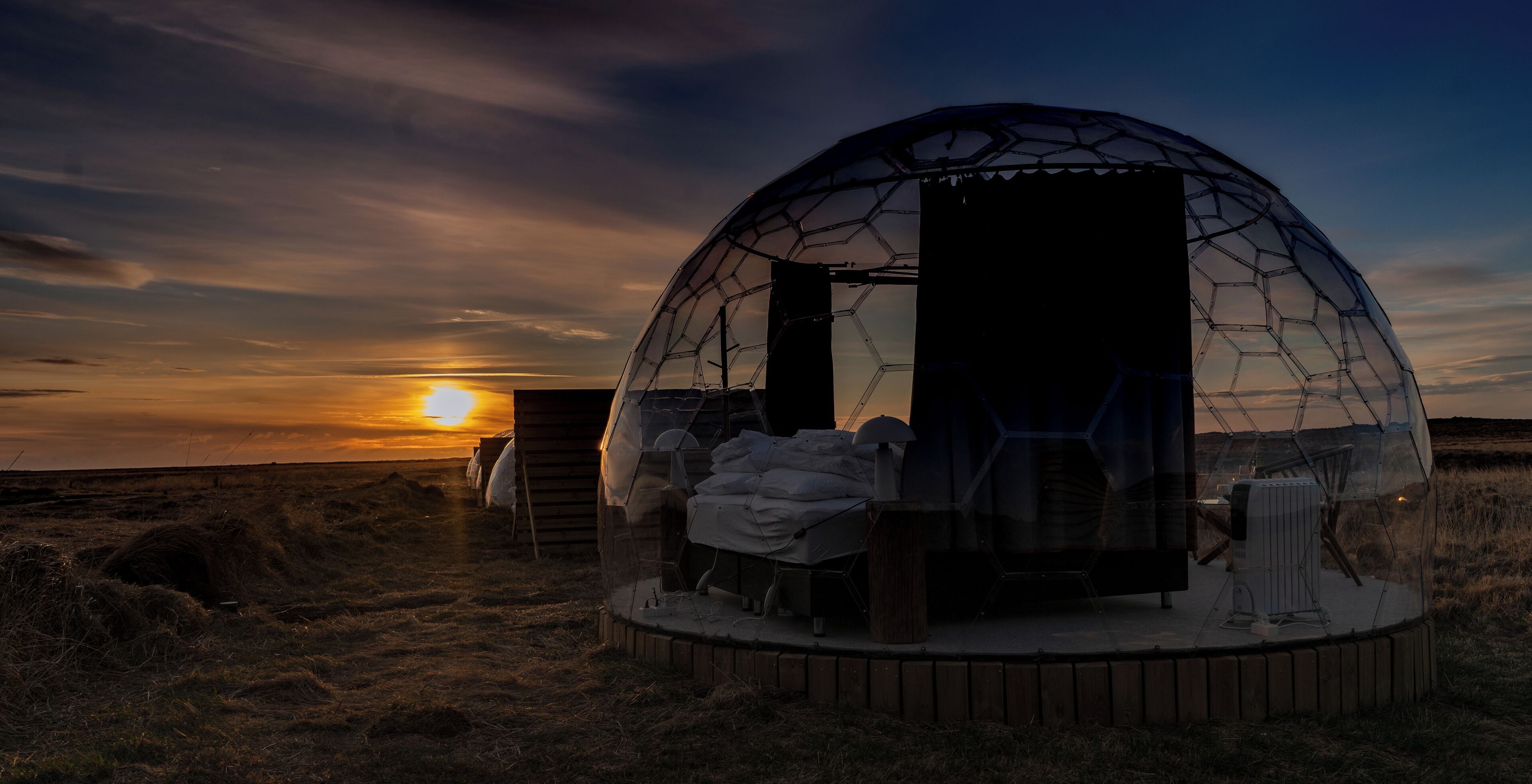 A geodesic dome structure with a wooden platform and seating area, set against a sunset backdrop.