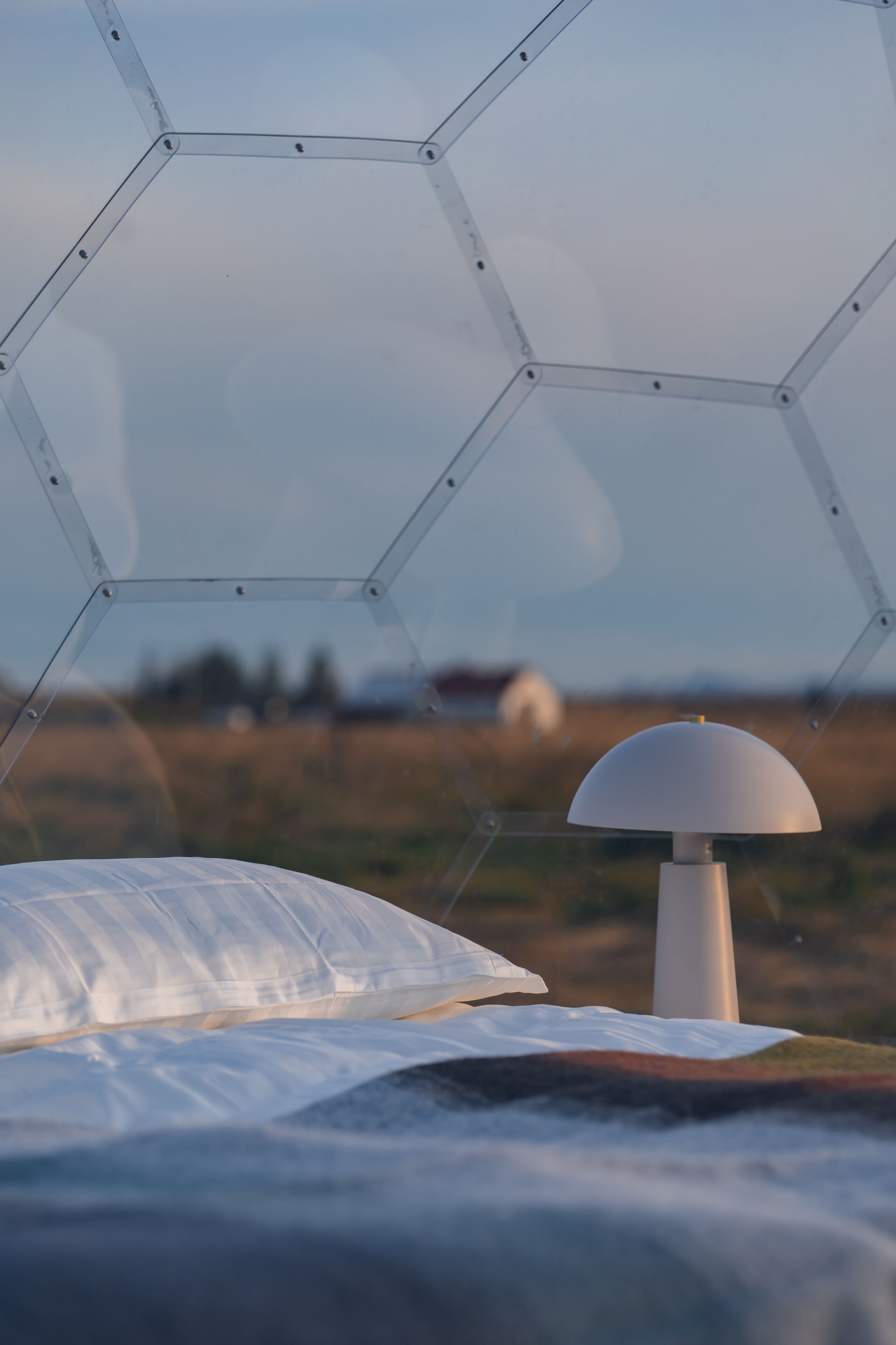 A bed with a white pillow and a white lamp in a room with a hexagonal window structure.
