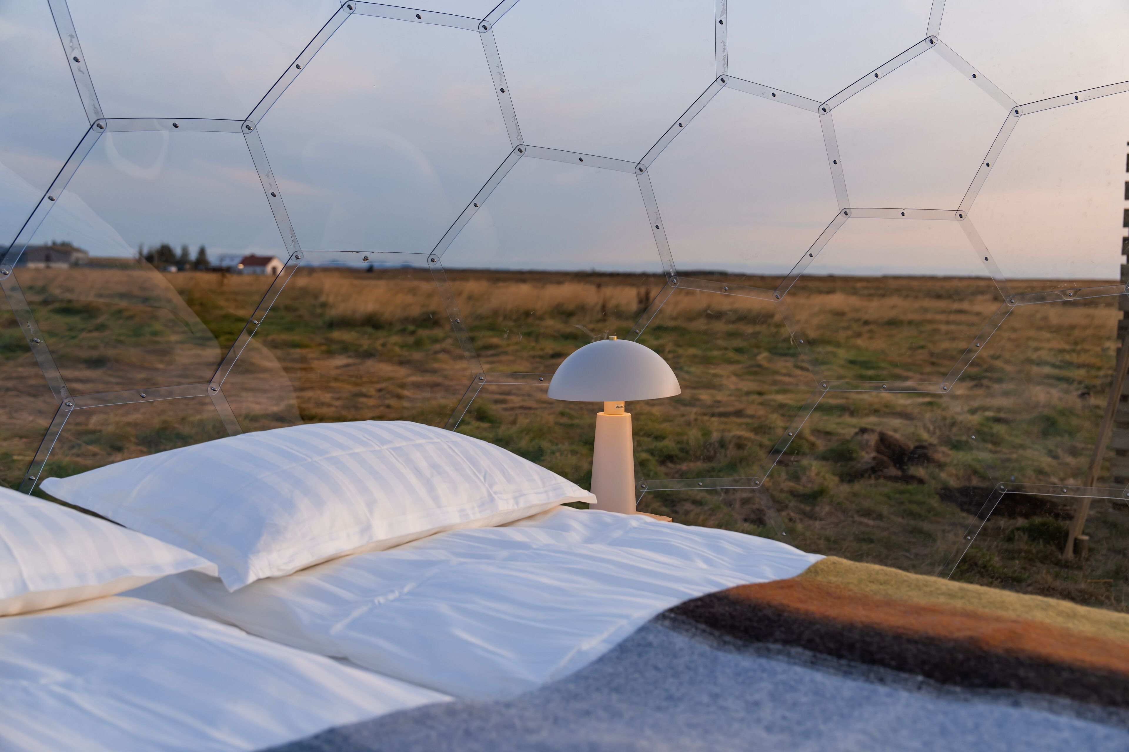 Een bed met wit beddengoed, een lamp en uitzicht op een grasveld door een geometrisch raam.
