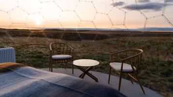 Een zeshoekige glazen constructie met twee houten stoelen en een kleine tafel in een open veld bij zonsondergang.