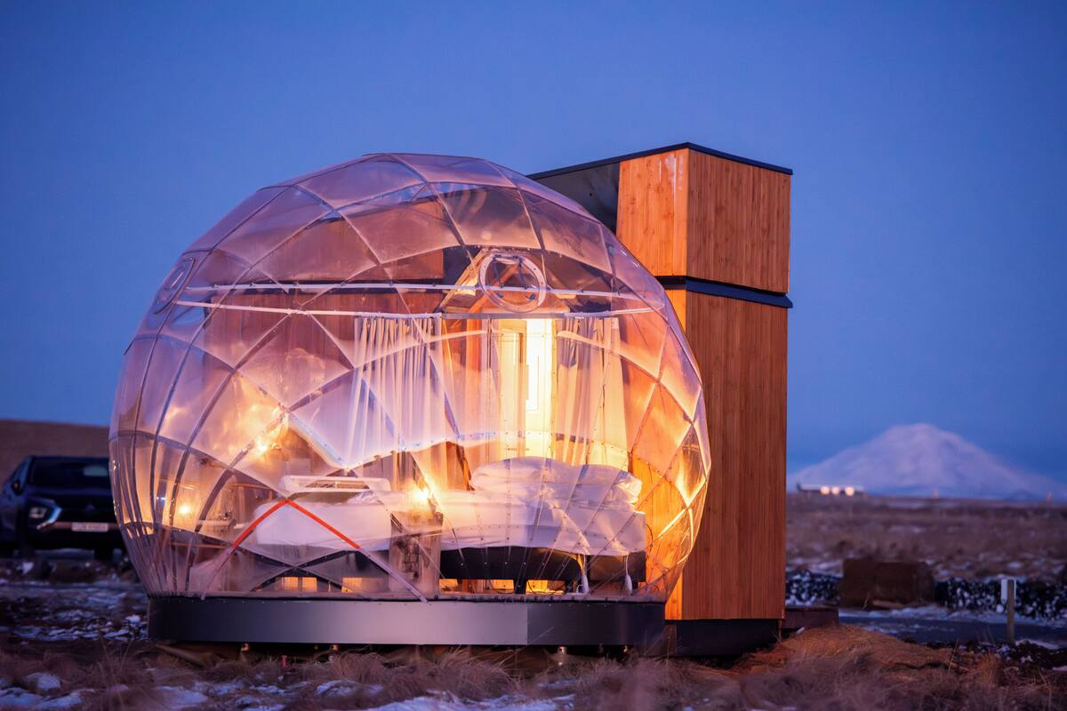 Eine transparente geodätische Kuppel mit Holzfassade, Innenraum mit Bett und Beleuchtung sowie ein Berg im Hintergrund.