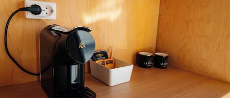 Uma cafeteira numa prateleira de madeira, com uma caixa branca contendo cápsulas de café e duas chávenas pretas.
