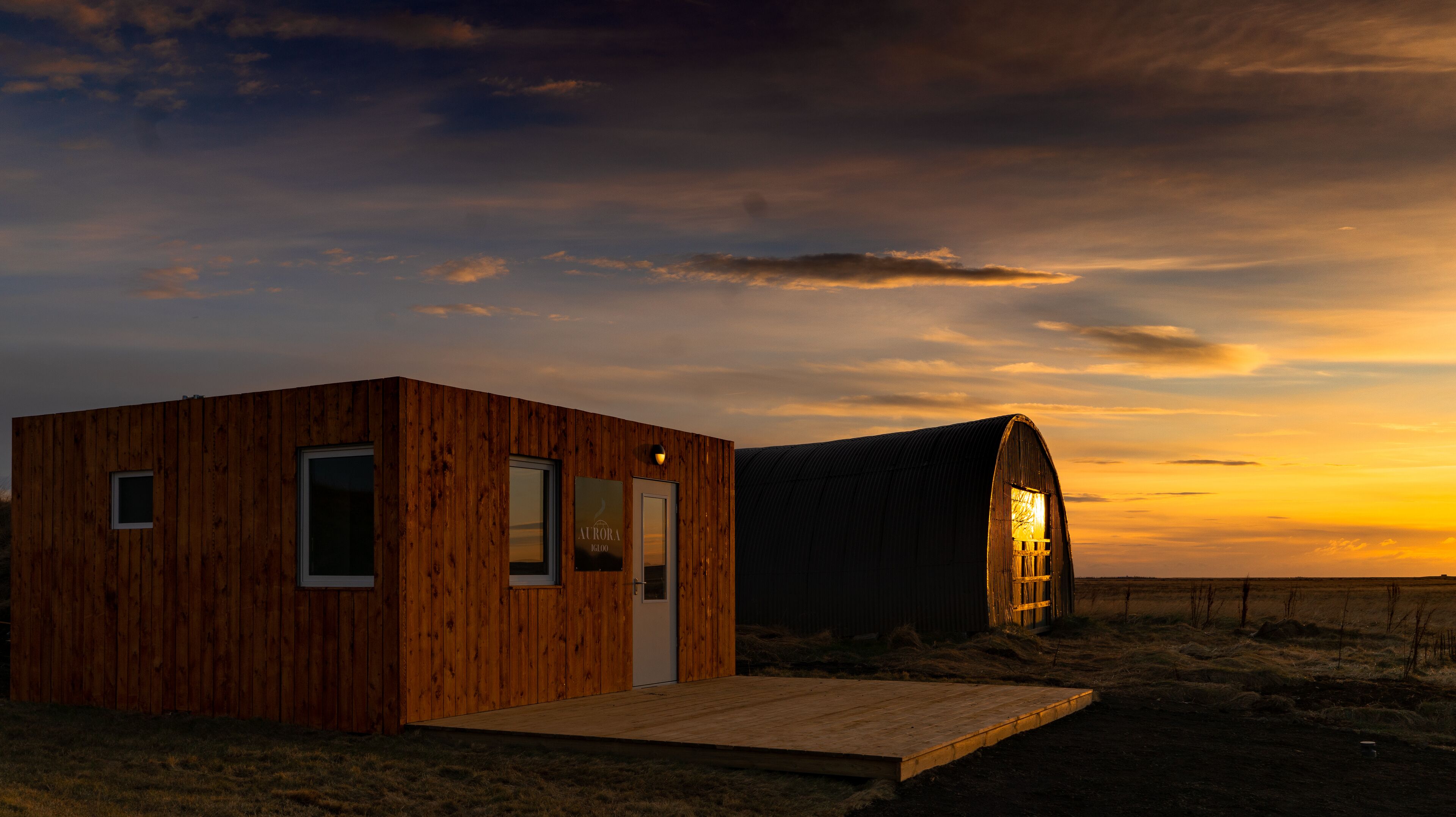 A wooden cabin with a door and windows, a wooden deck, and a barn with a light inside.