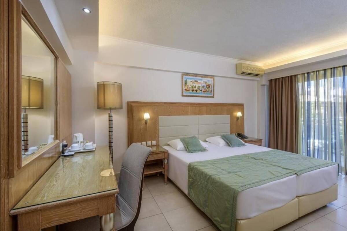 A hotel room with a bed, a desk, a chair, a mirror, a nightstand, and a window with curtains.