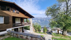 A two-story wooden house with a balcony, stone foundation, and a view of mountains.