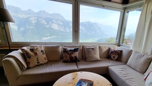 A beige sofa with decorative pillows and a round wooden coffee table in a room with large windows offering a mountain view.