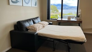 A bedroom with a bed, a black leather sofa, a wooden desk, and a large window with a view of a lake and mountains.