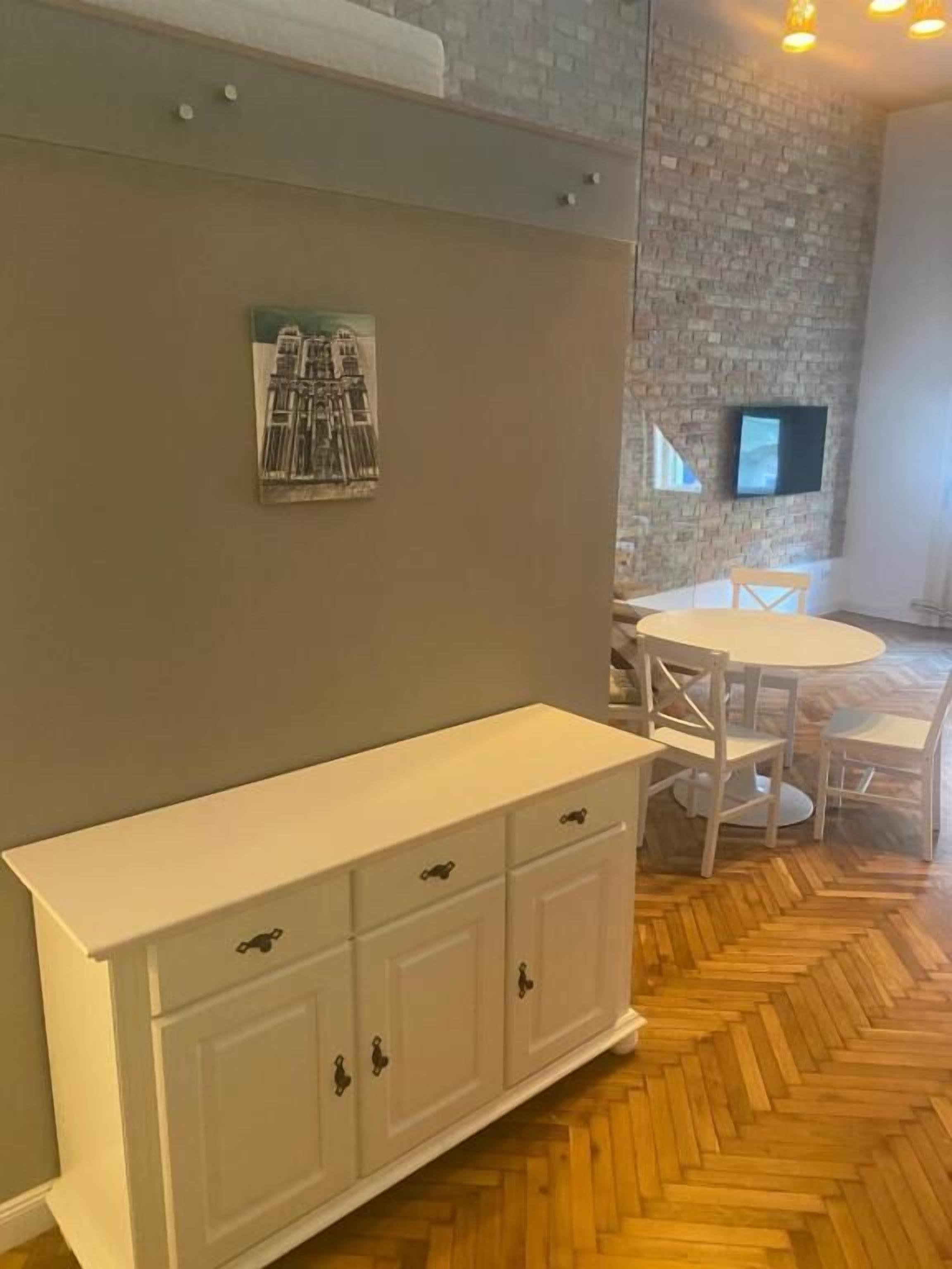A room with a herringbone wooden floor, a white cabinet with drawers, a round table with chairs, a flat-screen TV mounted on a brick wall, and a framed picture on the wall.