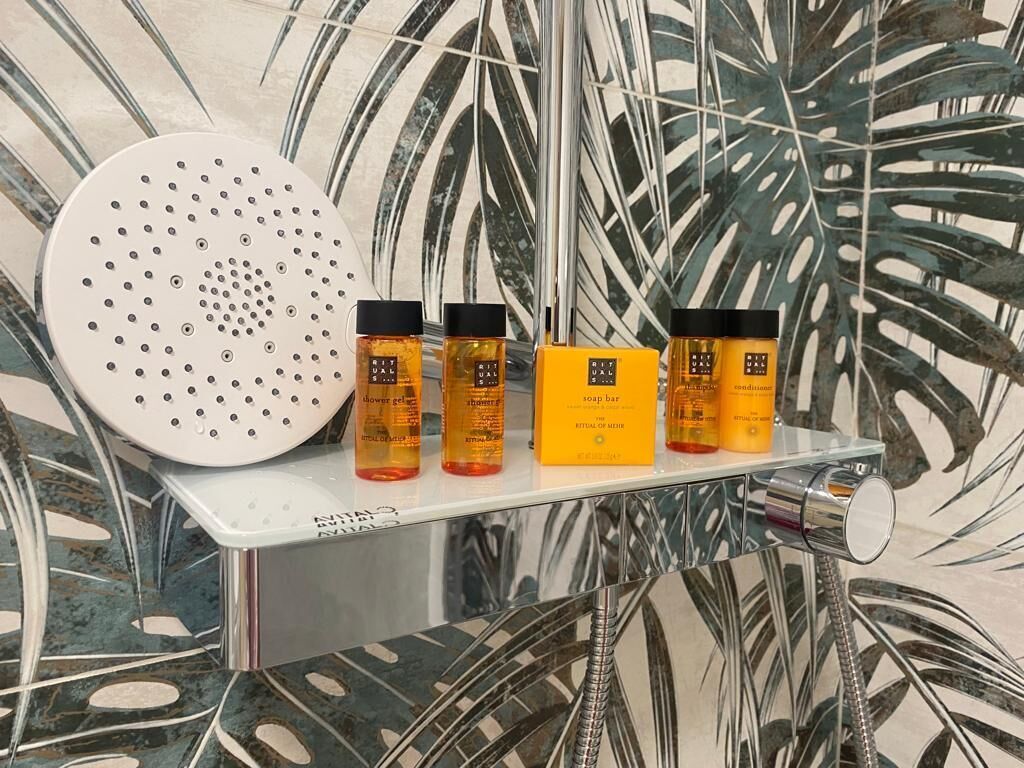 Un cabezal de ducha, cuatro botellas de jabón y una manguera de ducha sobre un estante frente a una pared con estampado de hojas tropicales.