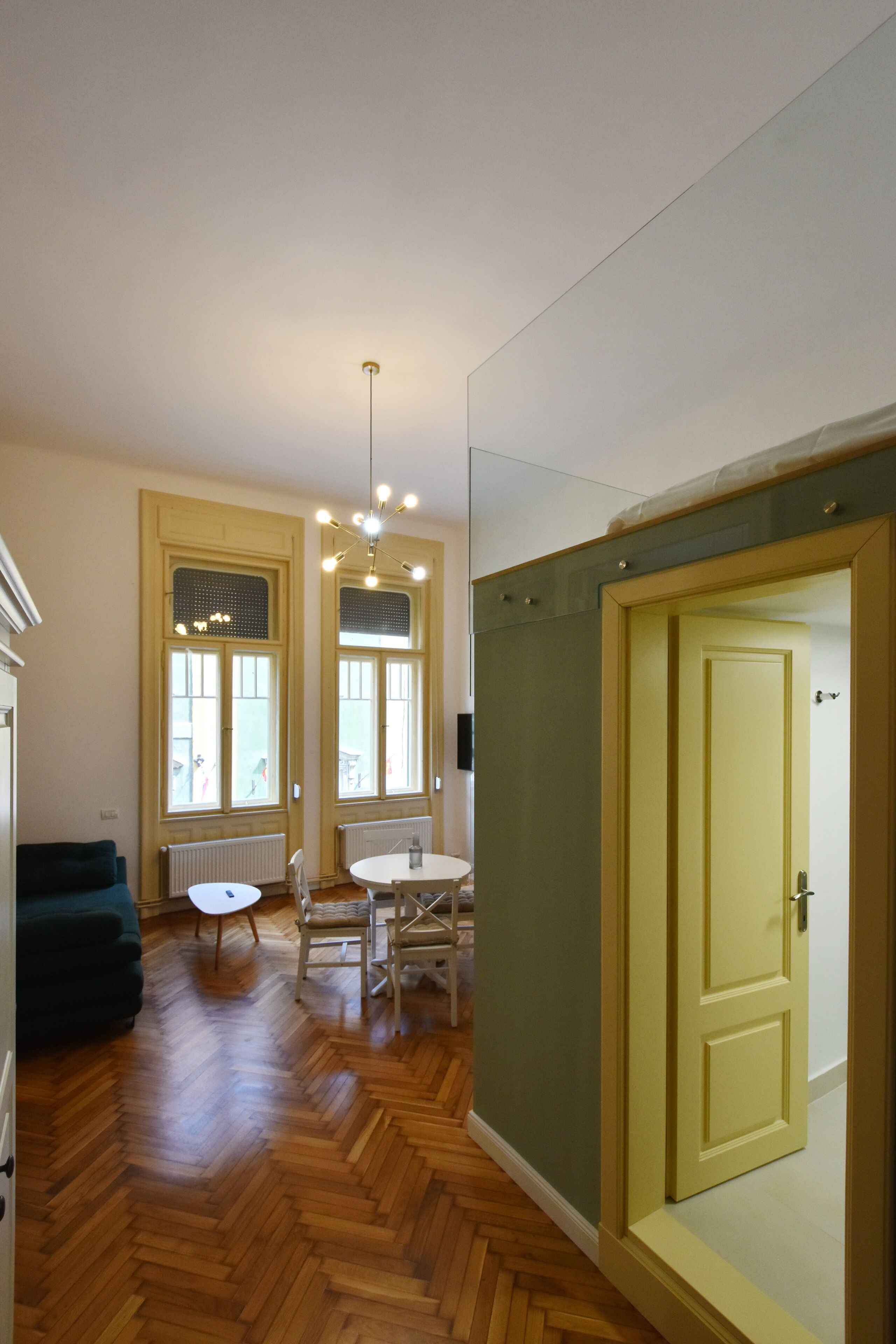 A room with a wooden herringbone floor, a green sofa, a white table with chairs, and a yellow door.