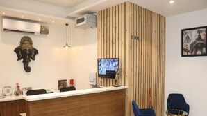 A reception area with a wooden reception desk, two blue chairs, a television, and a wall-mounted air conditioner. - Signum CityScapes The Royal Jewel (Hisar)