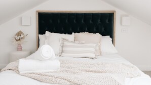 A neatly arranged bedroom with a dark upholstered headboard, white bedding, and a small bedside table with a vase of flowers. - Centrally Located Spacious & Luxurious Cottage (Parkes)