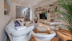 Un salon moderne avec un canapé, un pouf et des fauteuils, un téléviseur et une plante décorative.