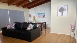 A living room with a black sofa, a wooden dining table, a white brick wall, and a framed picture of a palm tree. - Close to the Coast & Bellarine Peninsula (Moolap)