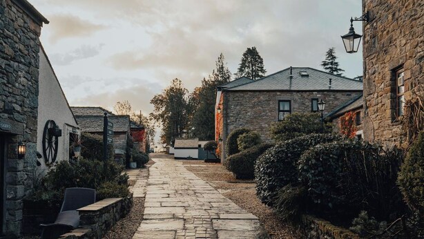 A cobblestone pathway leading through a rustic village with stone buildings, a sign for a café, and a lamp post.