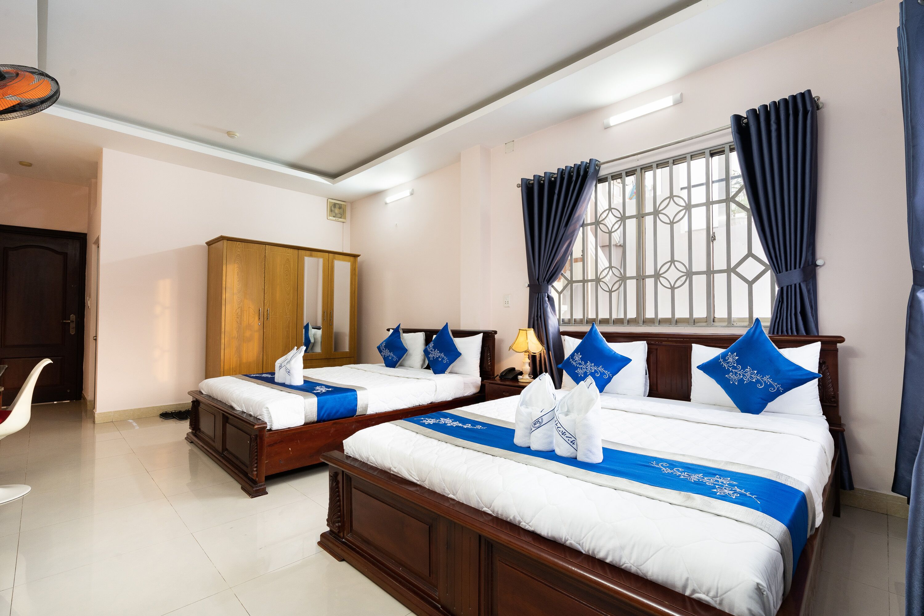 A hotel room with two beds, wooden headboards, white bedding, blue pillows, and a window with curtains.