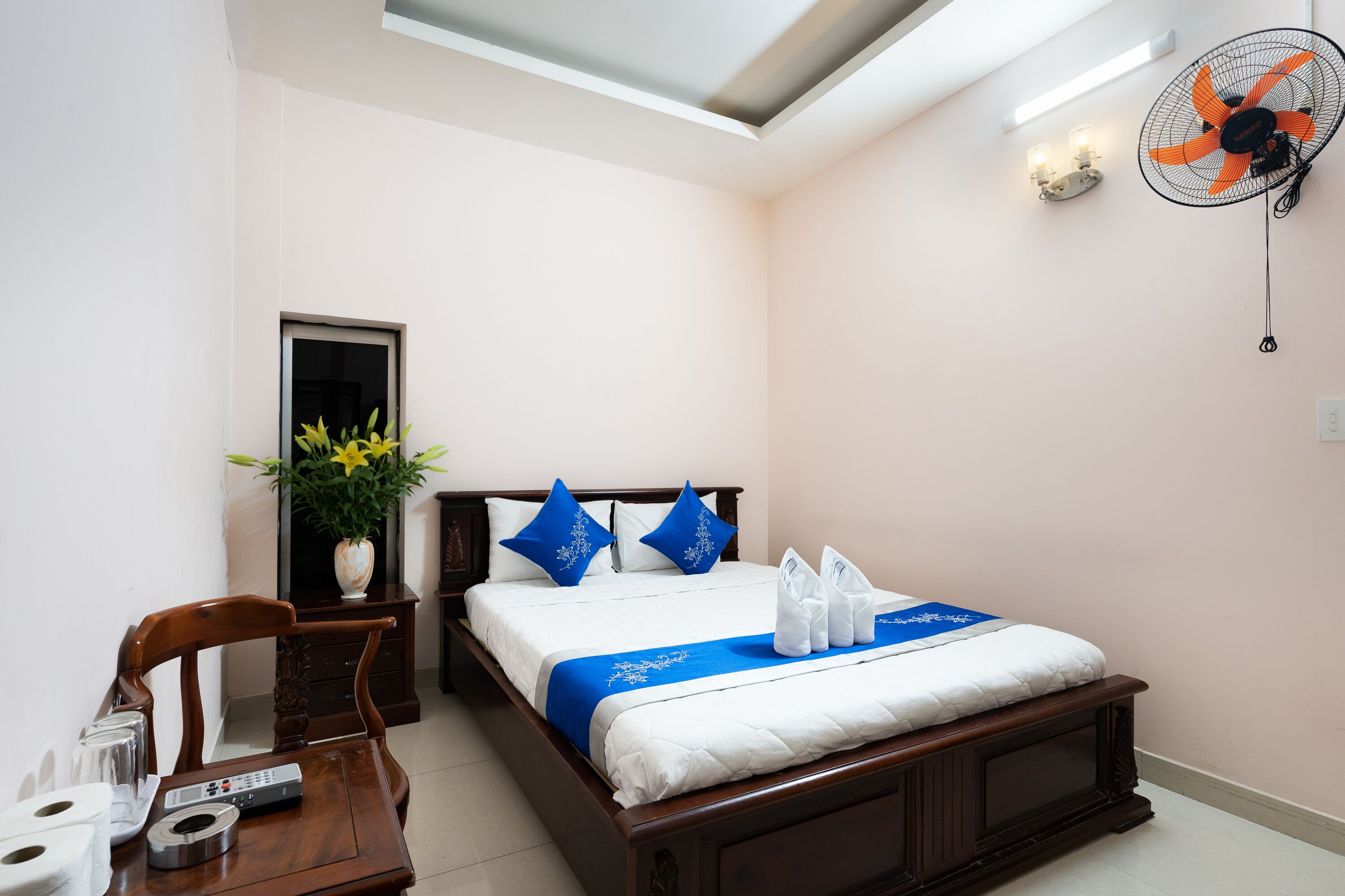 A hotel room with a bed, bedside table, chair, and a wall-mounted fan.