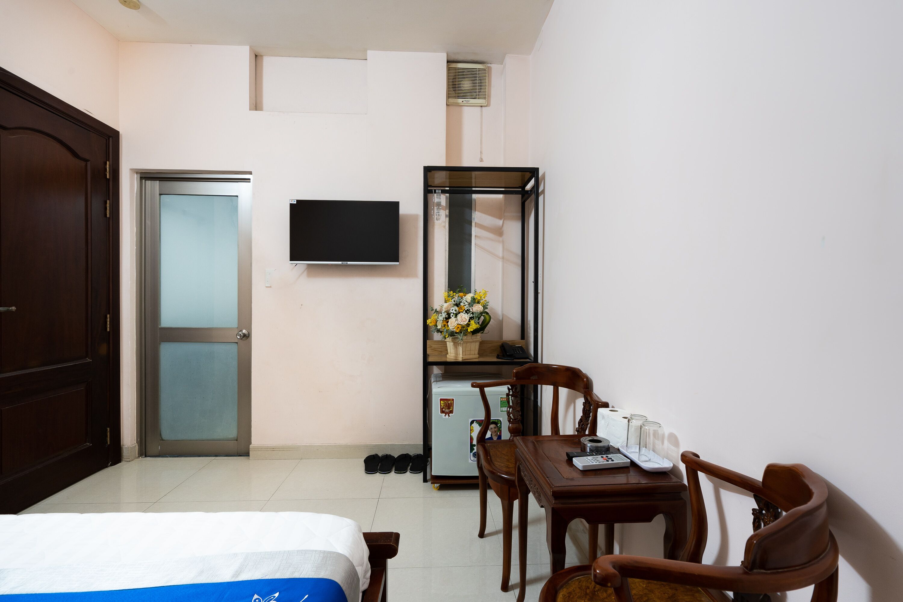 A hotel room with a bed, a desk with a chair, a refrigerator, a TV mounted on the wall, and a door.