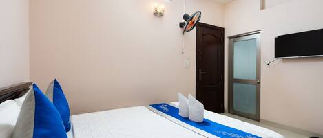 A neatly made bed with blue and white pillows, a blue runner, and a white blanket. There is a ceiling fan, a wall-mounted TV, and a door with a frosted glass panel.