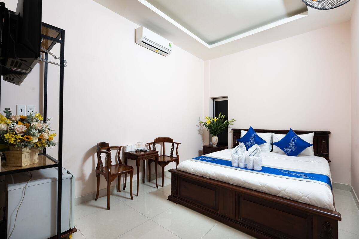A hotel room with a bed, a desk, two chairs, a small table, a potted plant, and an air conditioning unit.