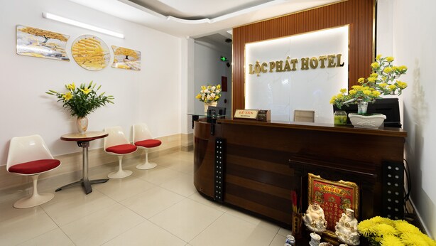 A hotel reception area with a wooden reception desk, a glass display case, and a reception sign. There are two chairs with red cushions and a small table with a vase of flowers. - Loc Phat Hotel (Ho Chi Minh City)