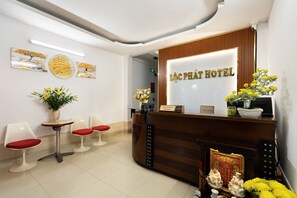 A hotel reception area with a wooden reception desk, a glass display case, and a reception sign. There are two chairs with red cushions and a small table with a vase of flowers. - Loc Phat Hotel (Ho Chi Minh City)