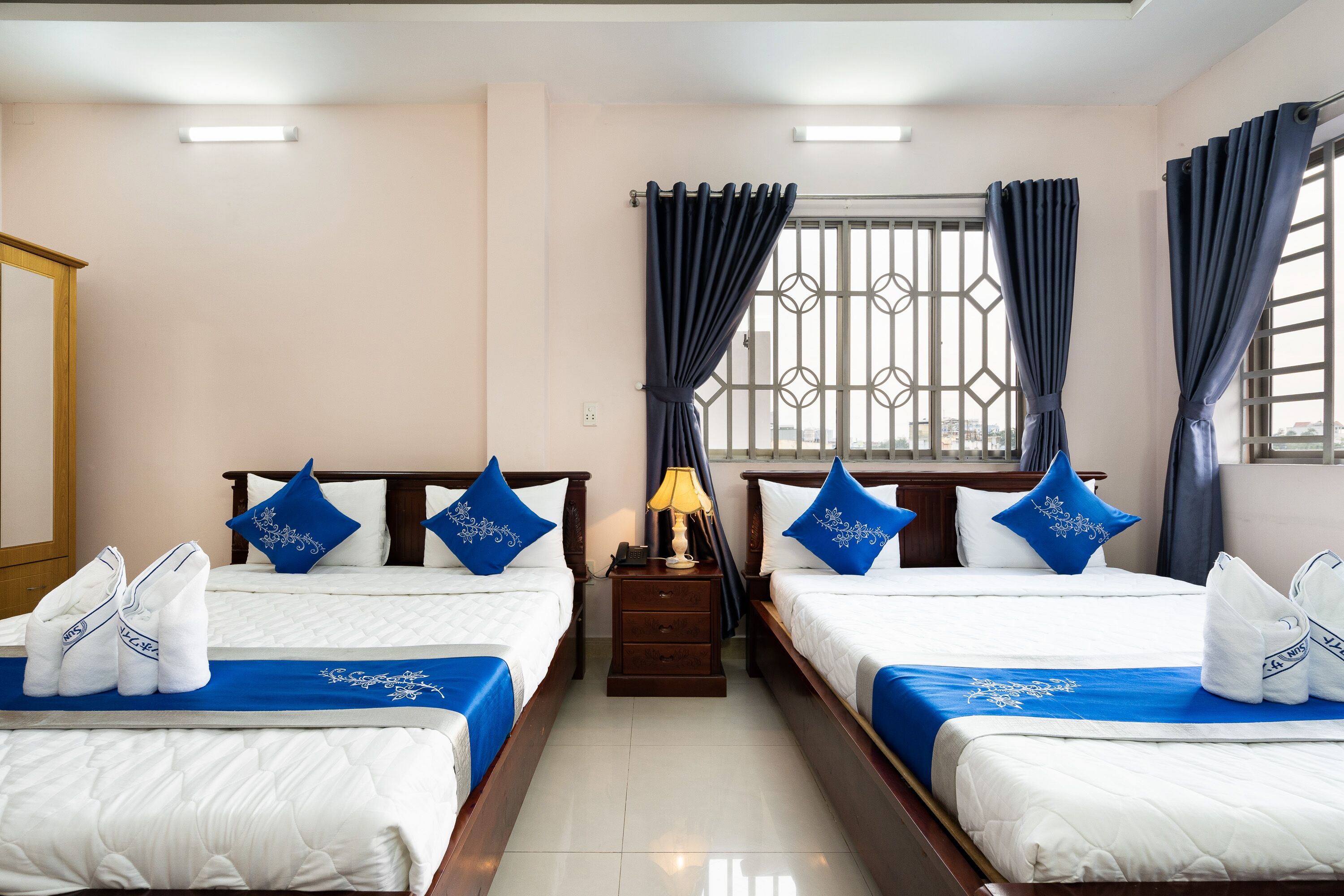 Habitación de hotel con dos camas, cada una con cubrecama y almohadas azules a juego, una mesita de noche con lámpara, y una ventana con celosías metálicas decorativas y cortinas.
