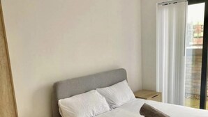 A neatly made bed with white linens and two pillows, a wooden nightstand with drawers, and a view of the outside through a window with curtains. - Salford City Retreat - Modern 2 Bedroom Apartment (Salford)