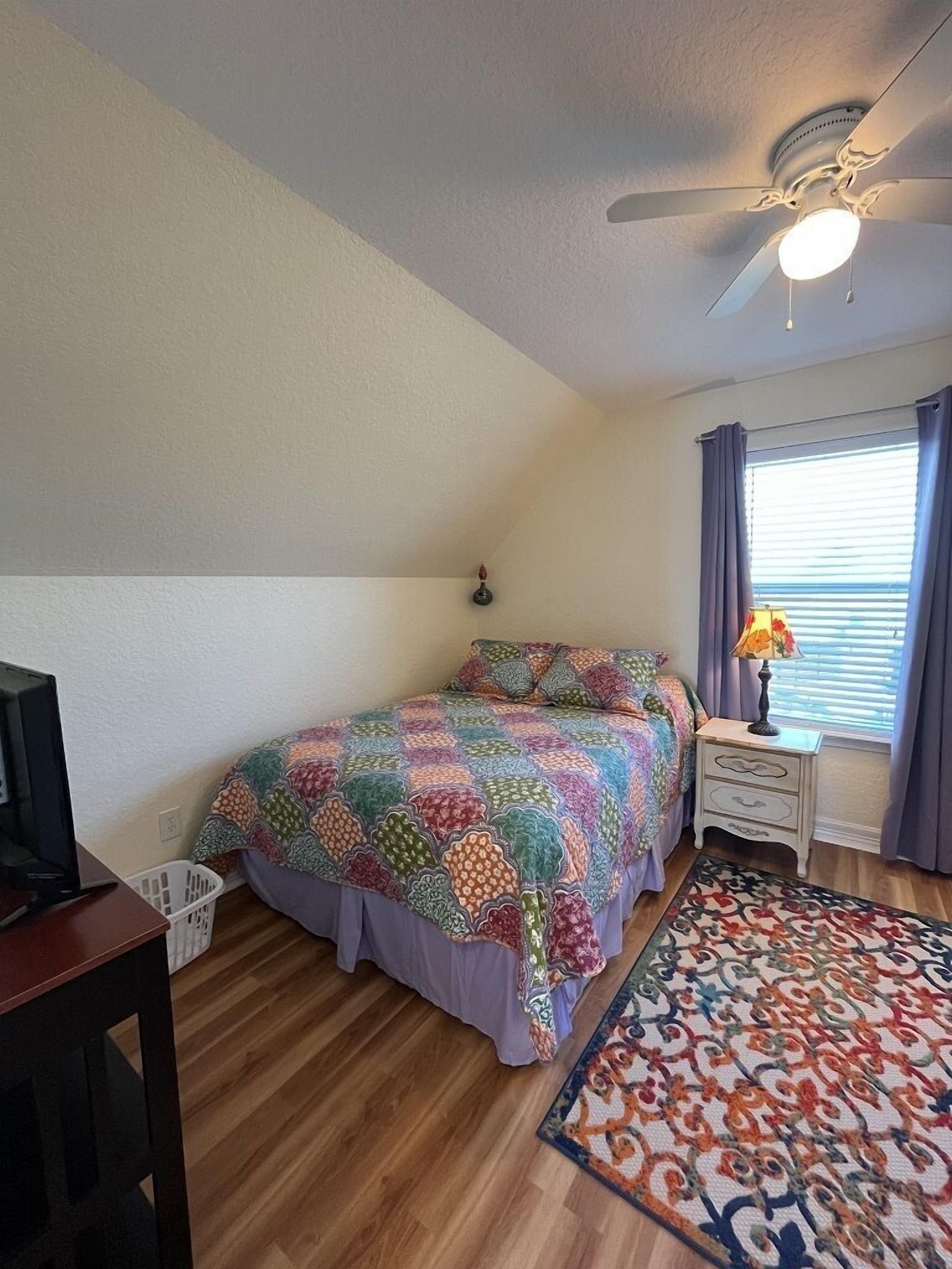 A bedroom with a bed, a nightstand, a lamp, a ceiling fan, a window with curtains, and a wooden floor.