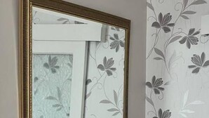 A room with a granite countertop, a wooden chair, a small table with a white vase of flowers, a wall-mounted mirror, and a floral wallpaper pattern. - Immaculate 3-bed House in Durham Near Sedgefield (Trimdon Station)