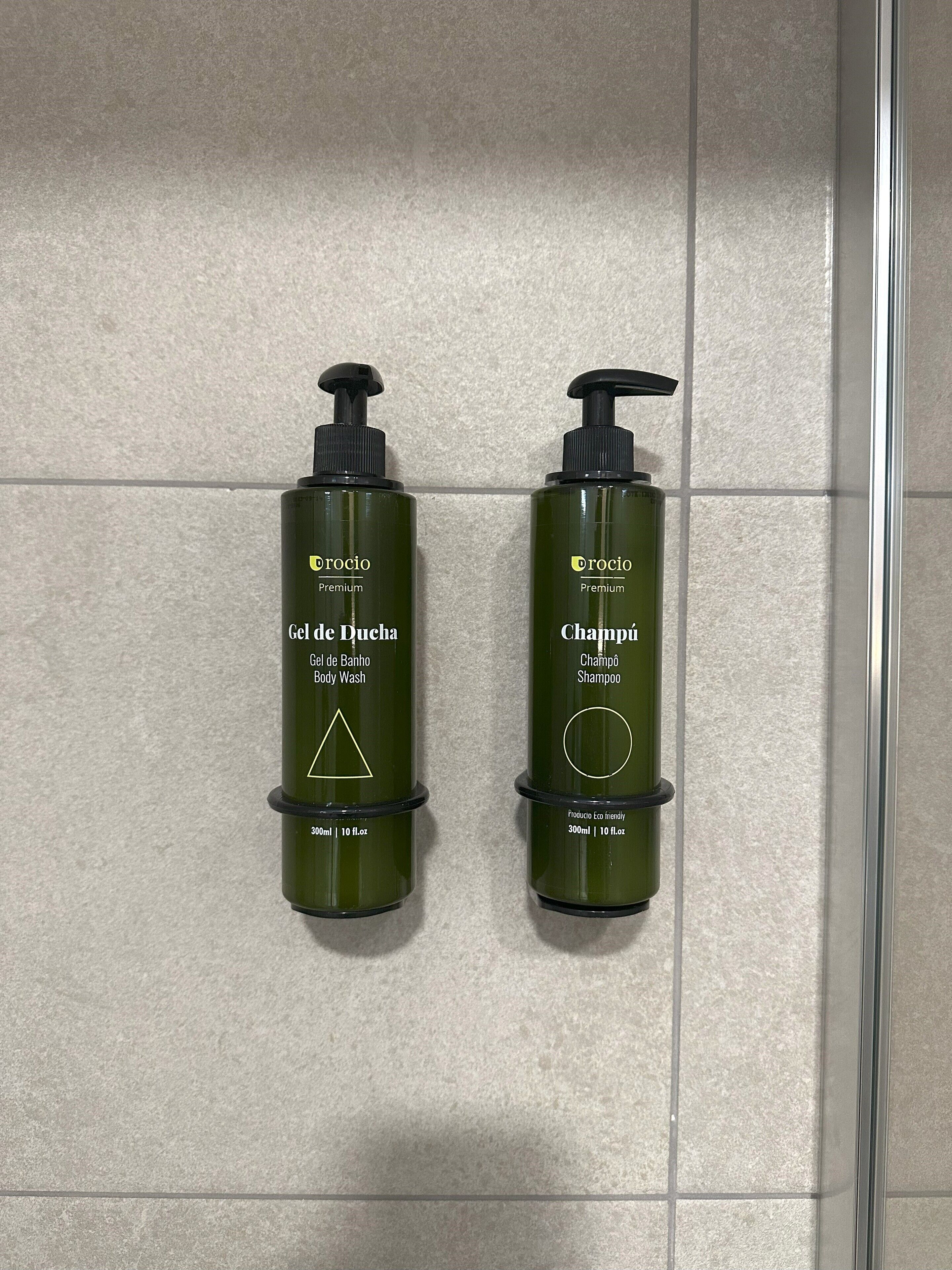 Deux bouteilles vertes de gel douche et de shampoing Drocio Premium sont posées sur un mur carrelé.