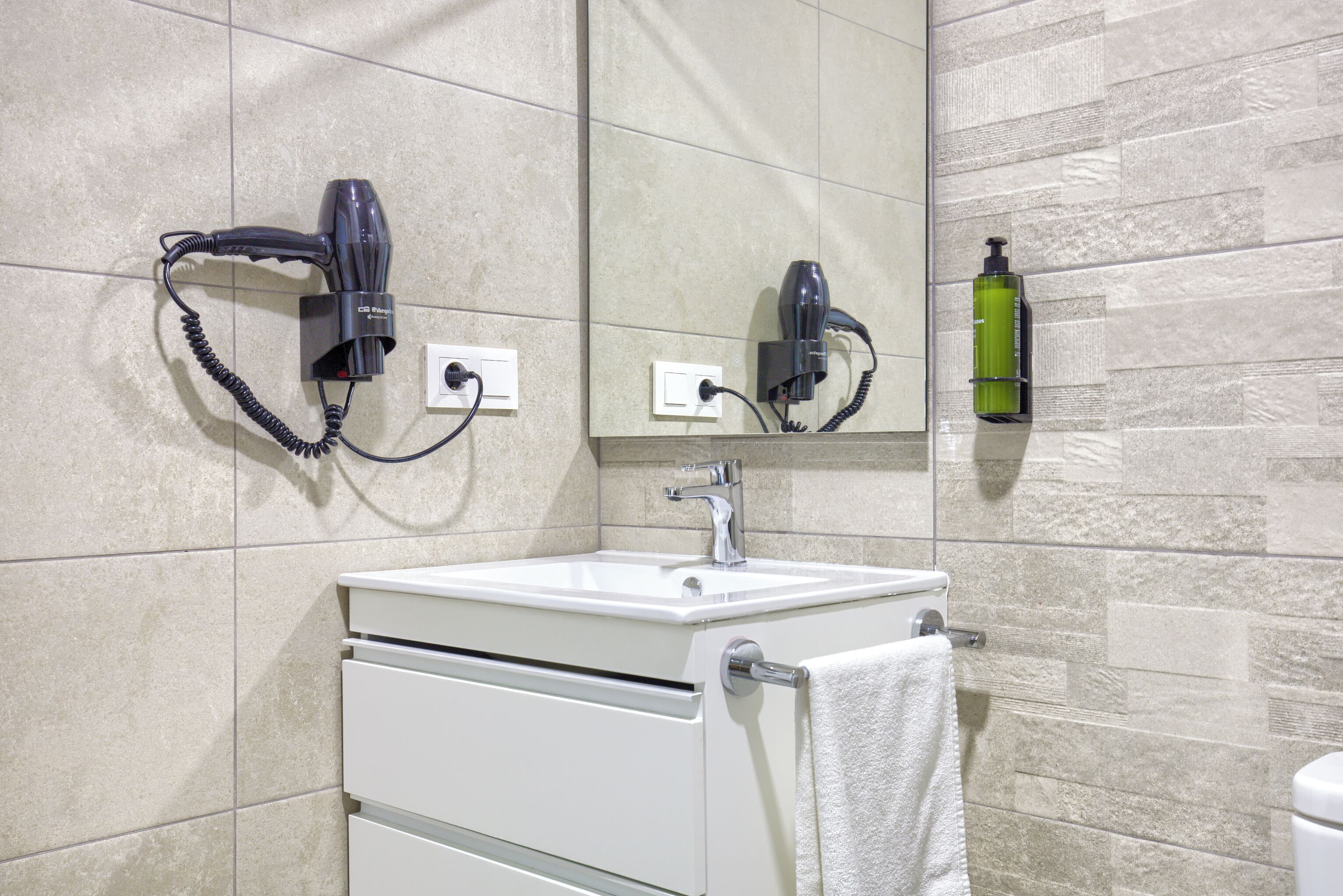 A bathroom with a sink, two hair dryers mounted on the wall, a mirror, and a green bottle of shampoo.
