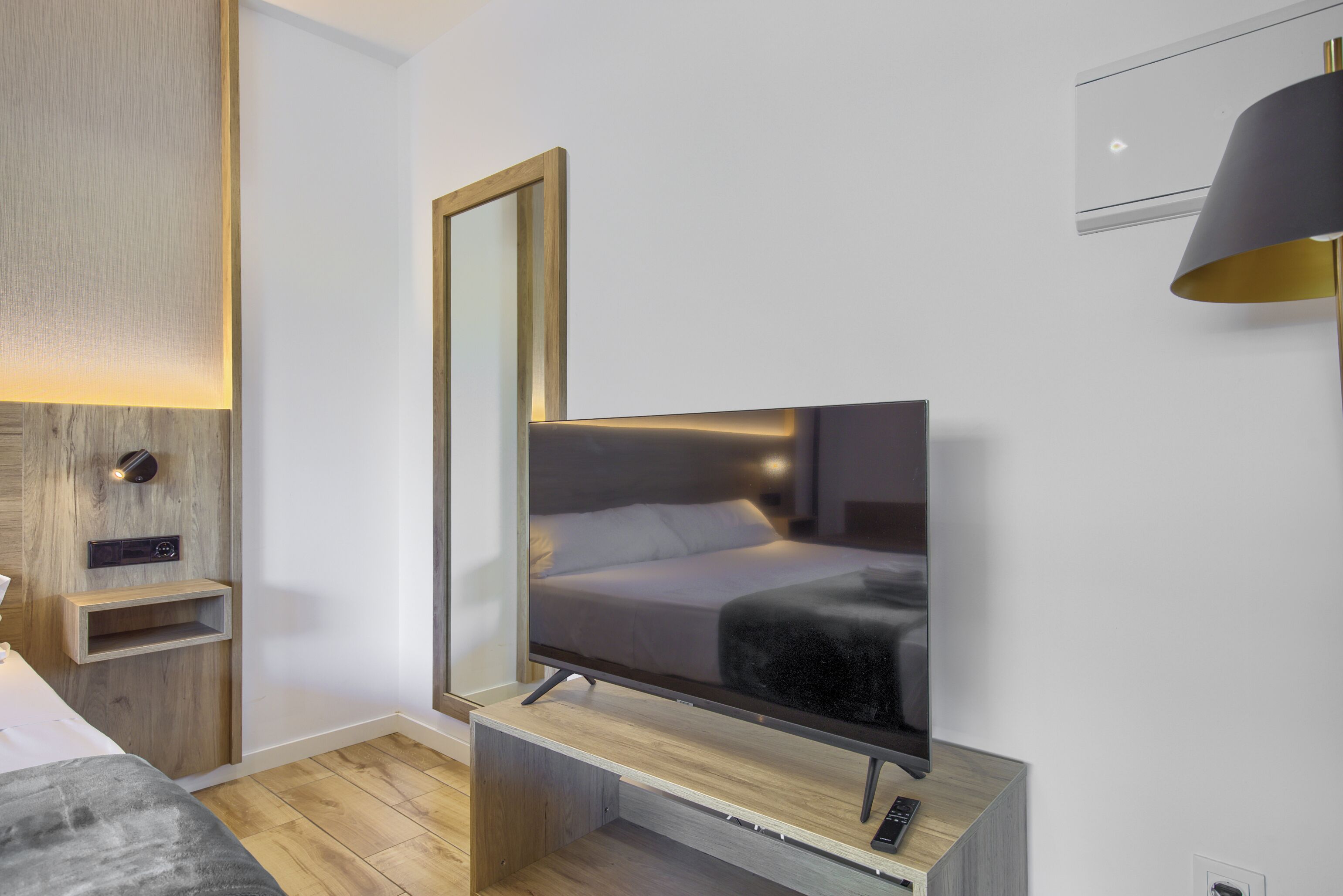 Una habitación de hotel moderna con una cama grande, un televisor de pantalla plana, una consola de madera, un espejo y una lámpara fijada a la pared.