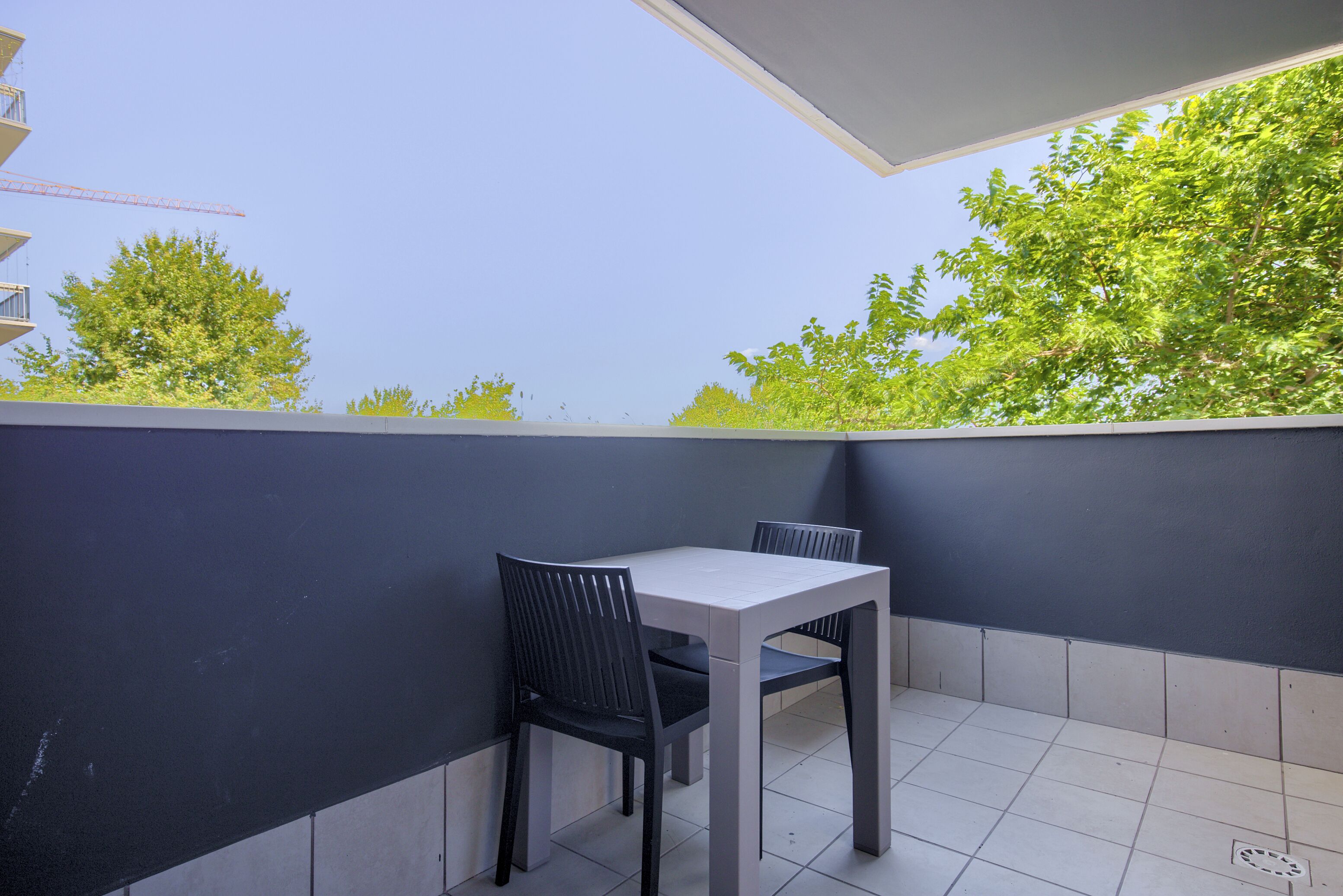 Un balcón con mesa y sillas, suelo de baldosas y vistas a árboles y un cielo despejado.