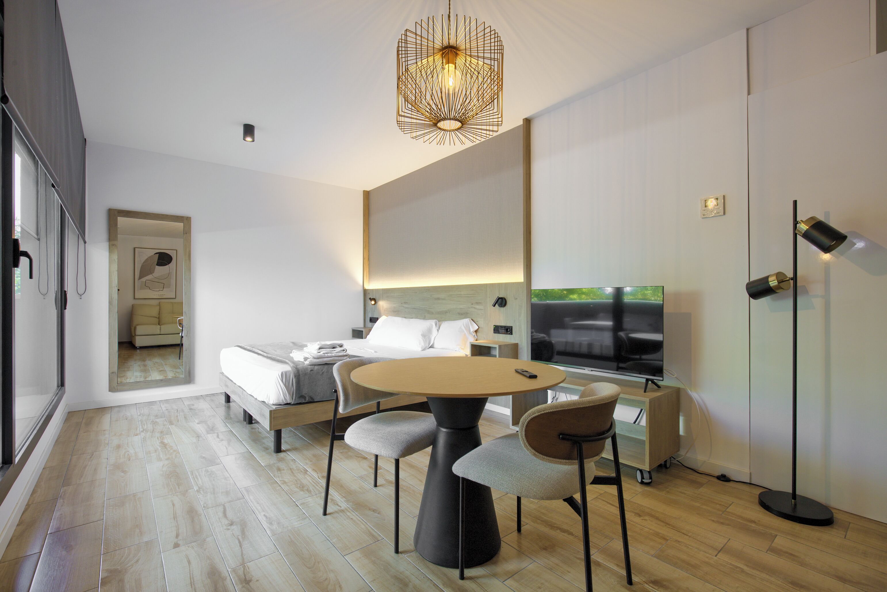 A modern hotel room with a round dining table, two chairs, a bed with white linens, a TV, and a large mirror.
