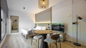 A modern hotel room with a round dining table, two chairs, a bed with white linens, a TV, and a large mirror.