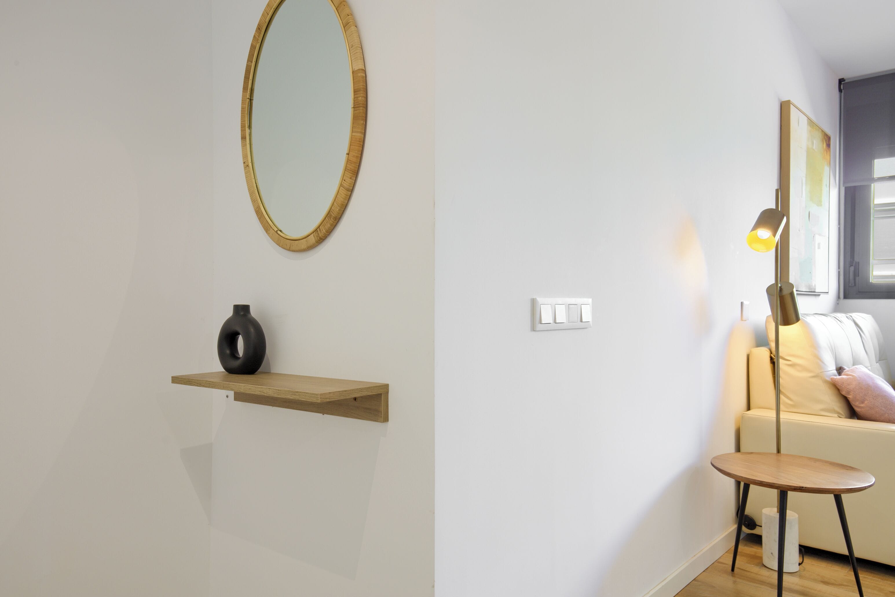 A modern living room with a round mirror, a small wooden shelf with a vase, a floor lamp, and a beige sofa.