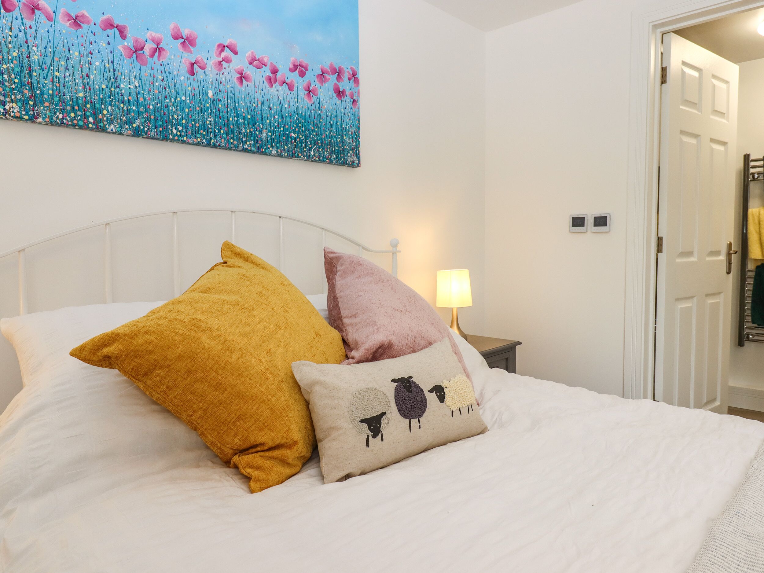 Ein ordentlich bezogenes Bett mit weißer Bettwäsche und drei Zierkissen, ein an der Wand hängendes Blumenbild und eine Tür zu einem Flur.