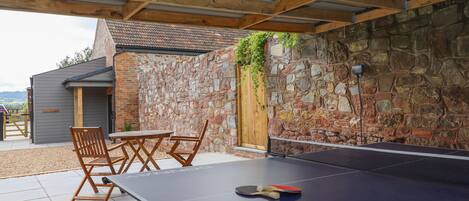 An outdoor ping pong table with chairs, a stone wall, and a wooden pergola.