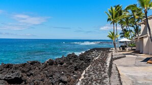 A coastal view with a rocky shoreline, palm trees, and a building with outdoor seating. - Summer Deal Oceanfront Kona Vacation Townhouse with Lanai and Sunsets (Kailua Kona)