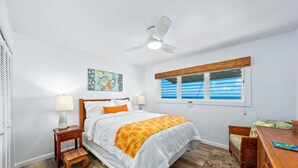 A bedroom with a bed, nightstands, a bedside table, a chair, a window with blinds, a ceiling fan, and a rug. - Summer Deal Oceanfront Kona Vacation Townhouse with Lanai and Sunsets (Kailua Kona)