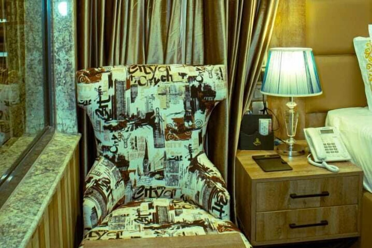 A small, cozy room with a bed, a bedside table with a lamp, and a chair with a patterned cushion.
