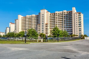 A large, multi-story hotel with a uniform facade, numerous balconies, and a parking area in the foreground.