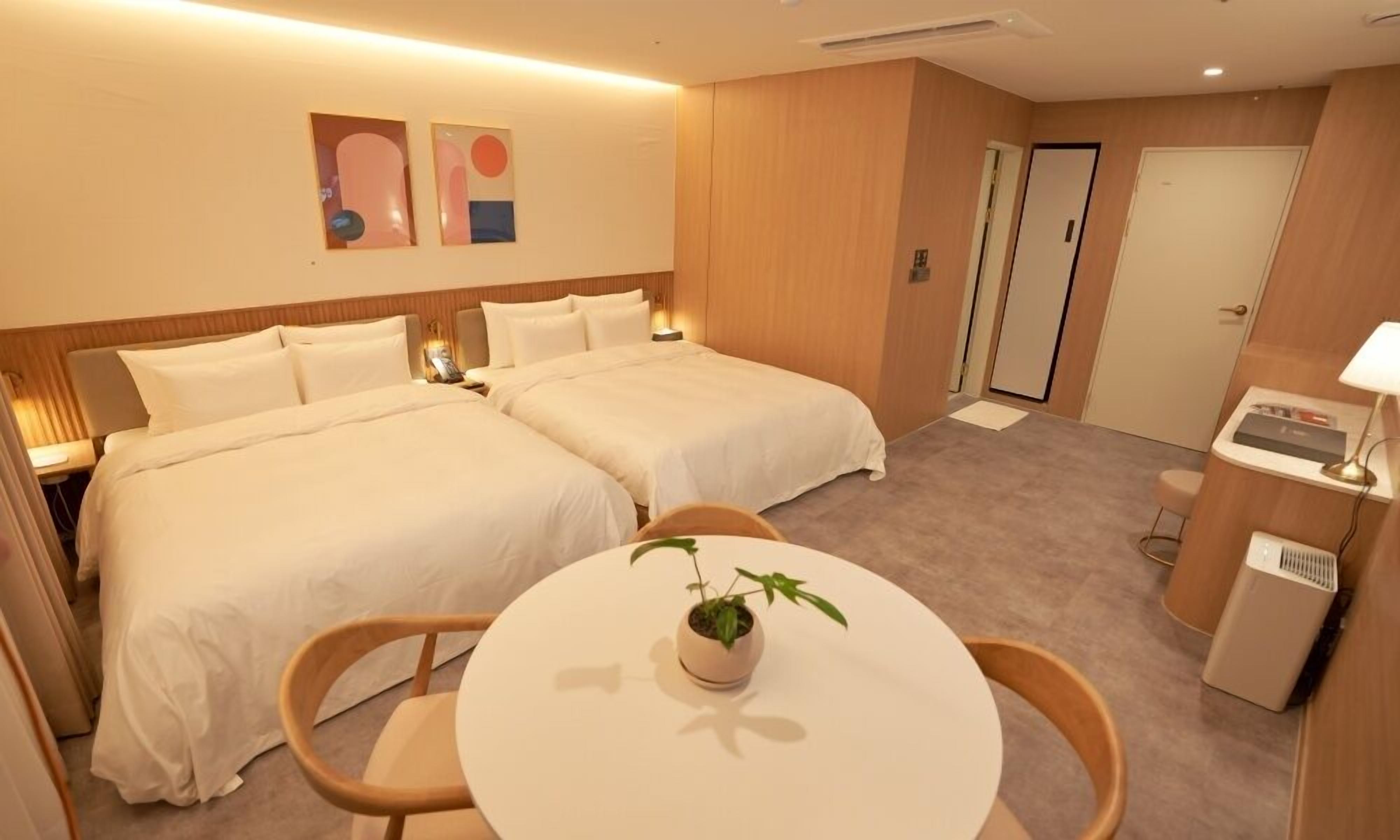 Quarto de hotel com duas camas, uma mesinha com uma planta, uma escrivaninha com abajur e uma porta.