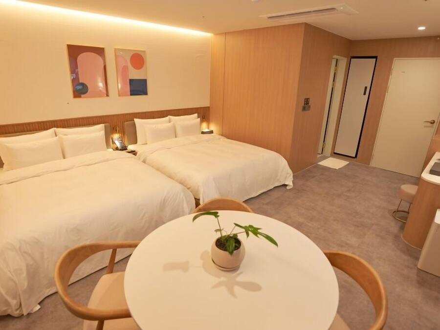 Quarto de hotel com duas camas, uma mesinha com uma planta, uma escrivaninha com abajur e uma porta.
