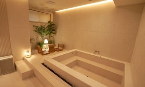 Un baño moderno con una bañera grande, una pequeña zona de estar con una silla y una mesa, y una planta en maceta.