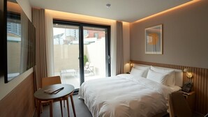 Habitación de hotel con cama, una mesita, una silla, una mesita de noche con lámpara y vistas al exterior por una puerta corredera.