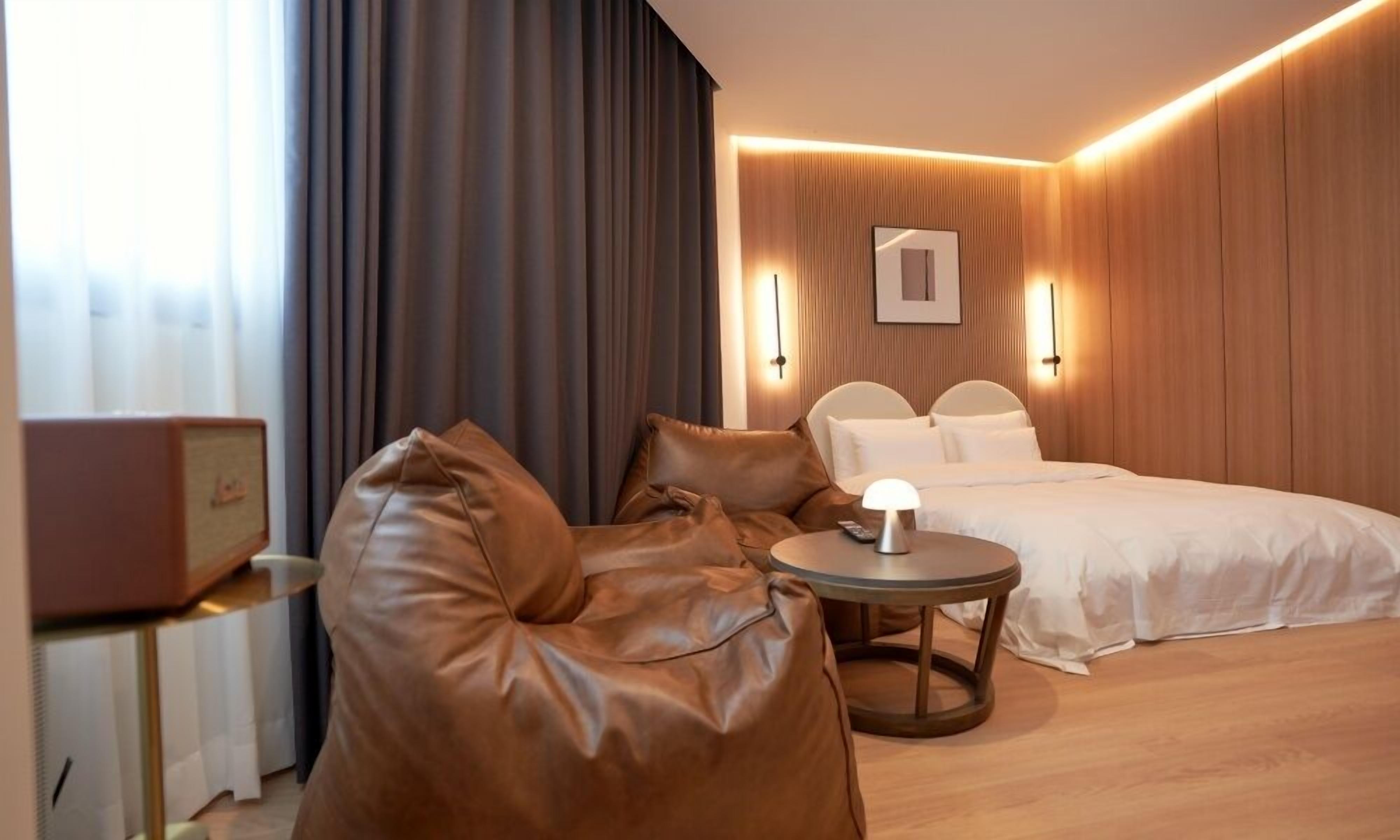 A modern hotel room with a leather armchair, a small round table, a bed with white bedding, and a wall-mounted light fixture.