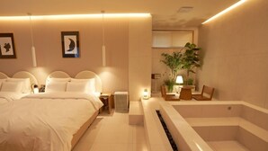 Habitación de hotel moderna con una cama grande, mesitas de noche, un escritorio pequeño, una silla, una planta y una bañera.